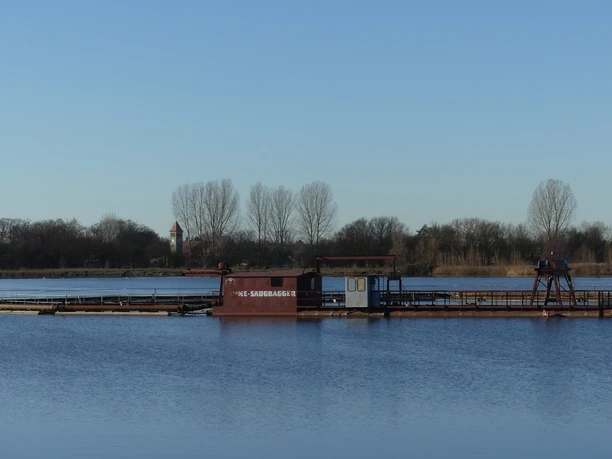 Baggersee zw. Anreppen und Delbrück Blauer See mit Saugbagger im Vordergrund, Ufer mit Bäumen und einem Kirchturm im Hintergrund.