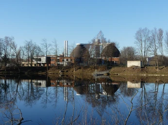 Blick auf die Kläranlage Kläranlage mit runden Tanks und Schornsteinen spiegelt sich in einem ruhigen Gewässer, umgeben von Bäumen.