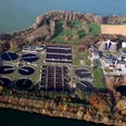 Luftaufnahme einer Kläranlage inmitten grüner Felder und Wälder mit Windkraftanlage im Hintergrund.