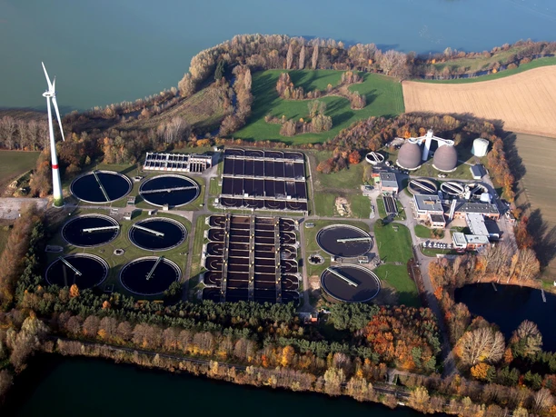 Luftaufnahme einer Kläranlage inmitten grüner Felder und Wälder mit Windkraftanlage im Hintergrund.