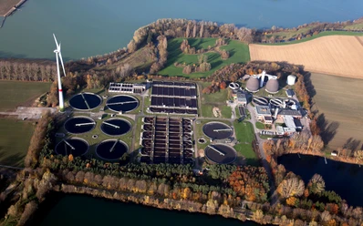 Luftaufnahme der Kläranlage Paderborn Luftaufnahme einer Kläranlage inmitten grüner Felder und Wälder mit Windkraftanlage im Hintergrund.