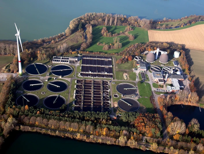 Luftaufnahme der Kläranlage Paderborn Luftaufnahme einer Kläranlage inmitten grüner Felder und Wälder mit Windkraftanlage im Hintergrund.