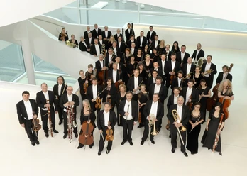 NWD_2013_1_c_sandra_kreutzer.jpg Eine große Gruppe von Musikern in formeller Abendkleidung posiert in einem modernen, hellen Raum.