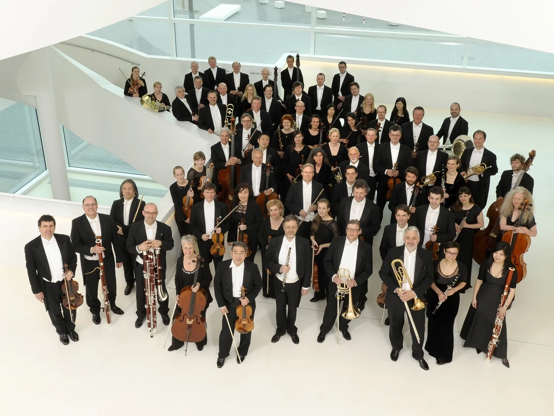 NWD_2013_1_c_sandra_kreutzer.jpg Eine große Gruppe von Musikern in formeller Abendkleidung posiert in einem modernen, hellen Raum.