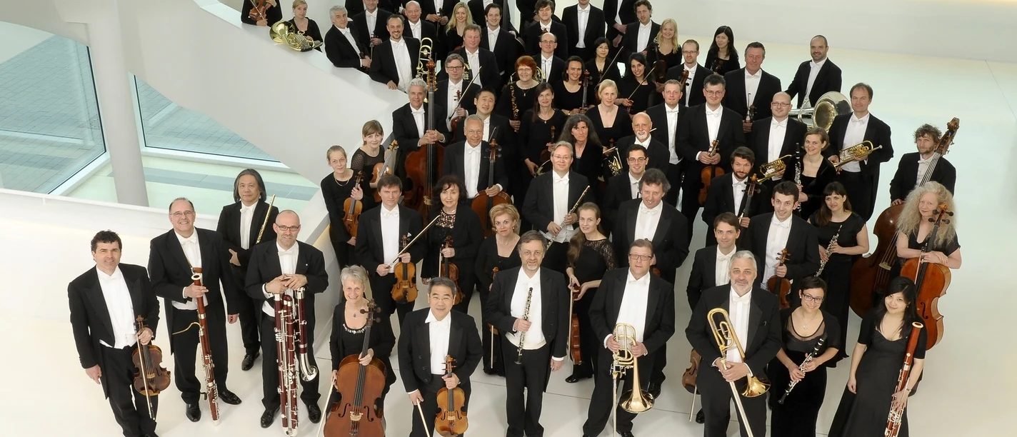 NWD_2013_1_c_sandra_kreutzer.jpg Eine große Gruppe von Musikern in formeller Abendkleidung posiert in einem modernen, hellen Raum.