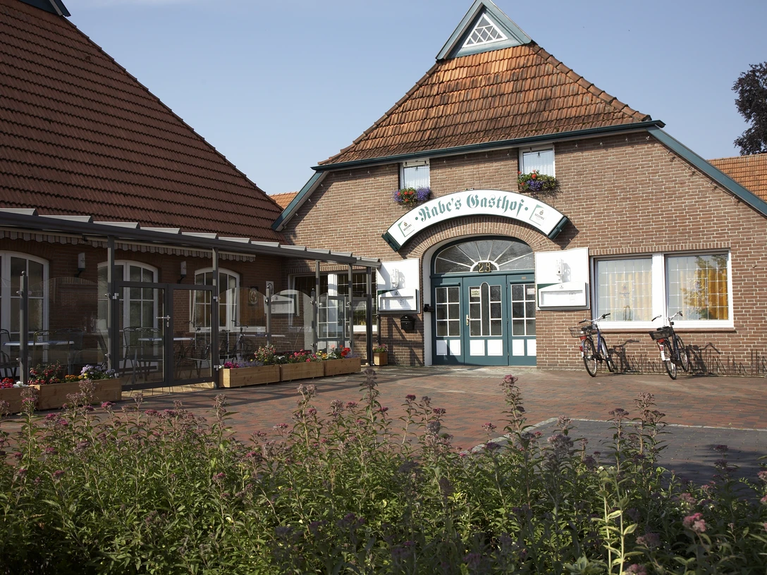Rabes Gasthof Das Bild zeigt Rabes Gasthof aus rotem Backstein mit einem großen Eingang und einem Ziegeldach. Vor dem Gasthof stehen zwei Fahrräder.