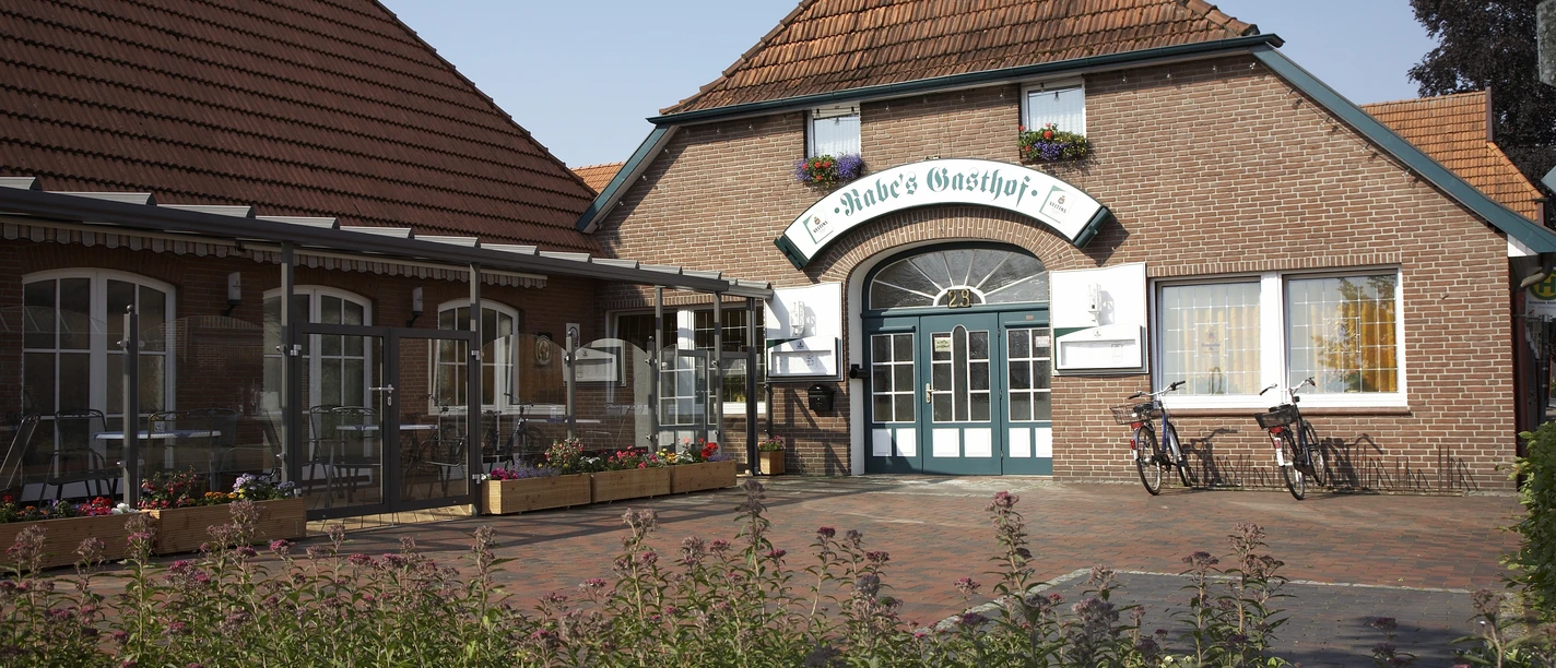 Rabes Gasthof Das Bild zeigt Rabes Gasthof aus rotem Backstein mit einem großen Eingang und einem Ziegeldach. Vor dem Gasthof stehen zwei Fahrräder.