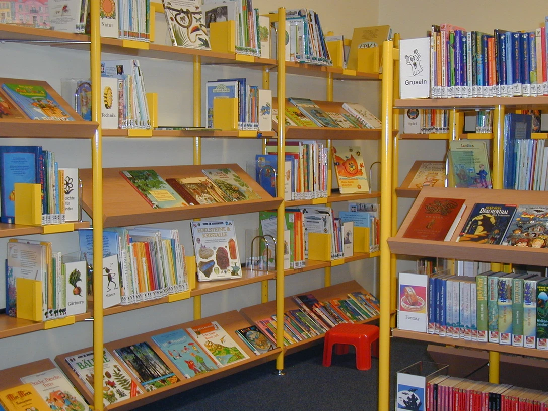 Kinderbücherei In der Kinderbücherei stehen bunte Bücher auf Regalen. Ein kleiner roter Hocker ist auf dem Boden sichtbar.