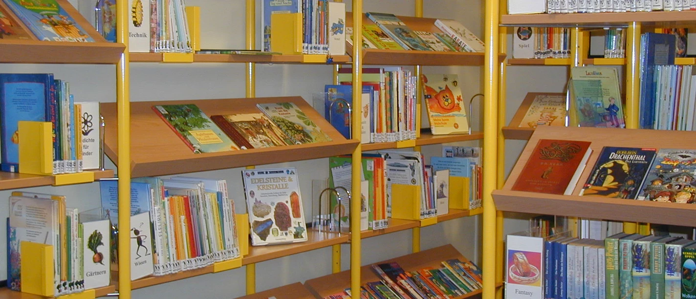 Kinderbücherei In der Kinderbücherei stehen bunte Bücher auf Regalen. Ein kleiner roter Hocker ist auf dem Boden sichtbar.