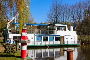 Kulturschiff ,,Blaue Donau" Weißes Kulturschiff mit blauen Elementen liegt am ruhigen Flussufer, umgeben von Bäumen und Sonne.