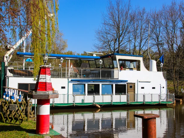 Kulturschiff ,,Blaue Donau" Weißes Kulturschiff mit blauen Elementen liegt am ruhigen Flussufer, umgeben von Bäumen und Sonne.