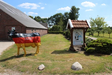 Milchtankstelle Hinrichs Das Bild zeigt eine Milchtankstelle auf einem Bauernhof. Eine auffällige Kuhfigur in schwarz, rot und gold, mit der Aufschrift "Die faire Milch", steht neben einem Informationsschild unter einem Holzdach. Im Hintergrund ist ein langes, rotes Backsteingebäude erkennbar sowie eine gepflegte Grasfläche mit einigen Bäumen.
