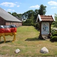 Milchtankstelle Hinrichs Das Bild zeigt eine Milchtankstelle auf einem Bauernhof. Eine auffällige Kuhfigur in schwarz, rot und gold, mit der Aufschrift "Die faire Milch", steht neben einem Informationsschild unter einem Holzdach. Im Hintergrund ist ein langes, rotes Backsteingebäude erkennbar sowie eine gepflegte Grasfläche mit einigen Bäumen.
