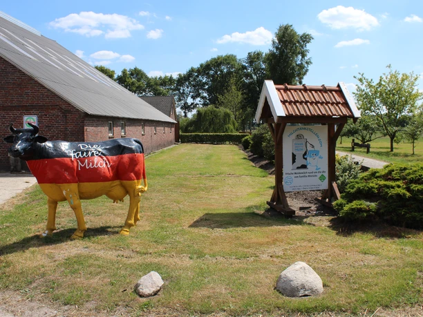 Milchtankstelle Hinrichs Das Bild zeigt eine Milchtankstelle auf einem Bauernhof. Eine auffällige Kuhfigur in schwarz, rot und gold, mit der Aufschrift "Die faire Milch", steht neben einem Informationsschild unter einem Holzdach. Im Hintergrund ist ein langes, rotes Backsteingebäude erkennbar sowie eine gepflegte Grasfläche mit einigen Bäumen.