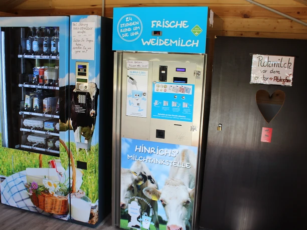 Milchtankstelle Hinrichs Im Inneren einer Holzhütte befinden sich drei Automaten. Der linke Automat bietet Getränke an. Der mittlere Automat ist als "Hinrichs Milchtankstelle" beschriftet und bietet frische Weidemilch an. Der rechte Automat hat eine herzförmige Öffnung, ergänzt um die Aufschrift "Rohmilch-bitte vor dem Verzehr abkochen"