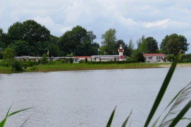 Ein idyllischer Campingplatz am Ufer eines Sees, geprägt von grüner Vegetation und einem markanten rot-weißen Leuchtturm im Hintergrund.