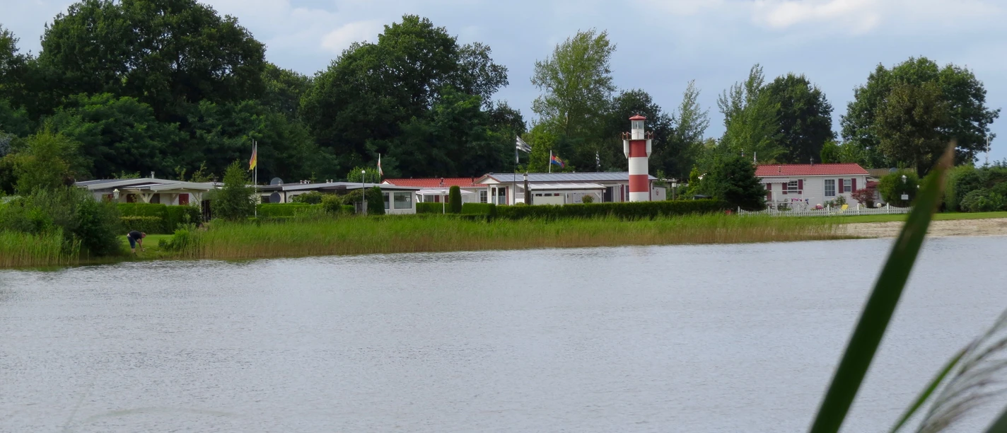 Campingplatz Nordloh Ein idyllischer Campingplatz am Ufer eines Sees, geprägt von grüner Vegetation und einem markanten rot-weißen Leuchtturm im Hintergrund.