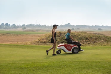 Zwei Frauen auf einem Golfplatz, eine geht mit Golfschläger, die andere sitzt in einem Golfmobil.