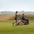 Zwei Frauen auf einem Golfplatz, eine geht mit Golfschläger, die andere sitzt in einem Golfmobil.