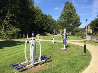 Outdoor-Fitnessgeräte auf einer grünen Wiese, umgeben von Bäumen, unter blauem Himmel.