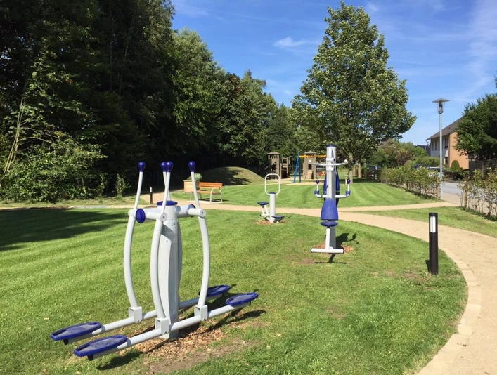 Generationenpark am Vogelgitter Outdoor-Fitnessgeräte auf einer grünen Wiese, umgeben von Bäumen, unter blauem Himmel.