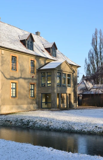 Historisches Gebäude mit schneebedecktem Dach, umgeben von einem zugefrorenen Wassergraben und Bäumen.