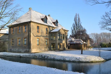 Bürgerbegegnungsstätte Haus Werther Historisches Gebäude mit schneebedecktem Dach, umgeben von einem zugefrorenen Wassergraben und Bäumen.