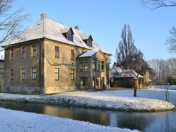 Bürgerbegegnungsstätte Haus Werther Historisches Gebäude mit schneebedecktem Dach, umgeben von einem zugefrorenen Wassergraben und Bäumen.