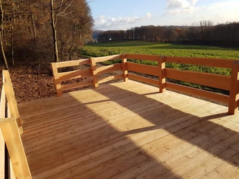 Rastplatz am Luisenturm Weg Holzplattform am Waldrand mit Blick auf grüne Wiesen und bewölkten Himmel in der Ferne.