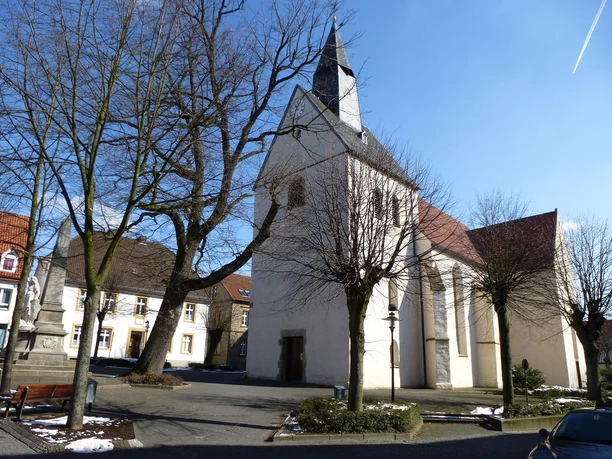 Eine historische Kirche mit weißem Turm, umgeben von kahlen Bäumen auf einem sonnigen Platz.