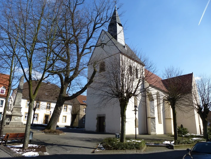 Mediendatei Eine historische Kirche mit weißem Turm, umgeben von kahlen Bäumen auf einem sonnigen Platz.