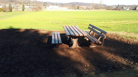 Rastplatz am Barenbergweg Holztisch mit zwei Bänken steht in ländlicher Umgebung, von Feldern und sanften Hügeln umgeben.