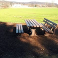 Rastplatz am Barenbergweg Holztisch mit zwei Bänken steht in ländlicher Umgebung, von Feldern und sanften Hügeln umgeben.