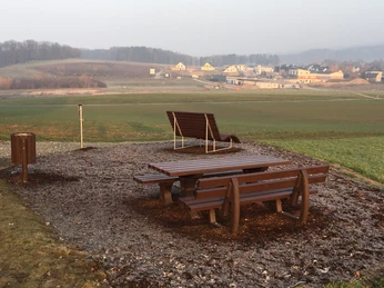 Holzpicknicktisch und Bank auf ruhigem Feldweg bei dunstigem Wetter mit Dorf im Hintergrund.