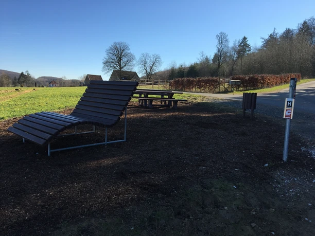 Rastplatz am Barenbergweg Eine Holzliege neben einem Tisch und Bänken auf einem Rastplatz am Feldrand, umgeben von Bäumen.