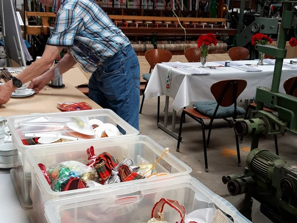 Vorbereitungen zum Webkurs Ein Mann arbeitet an einem Tisch in einer Werkstatt voller Bänder und textiler Maschinen.