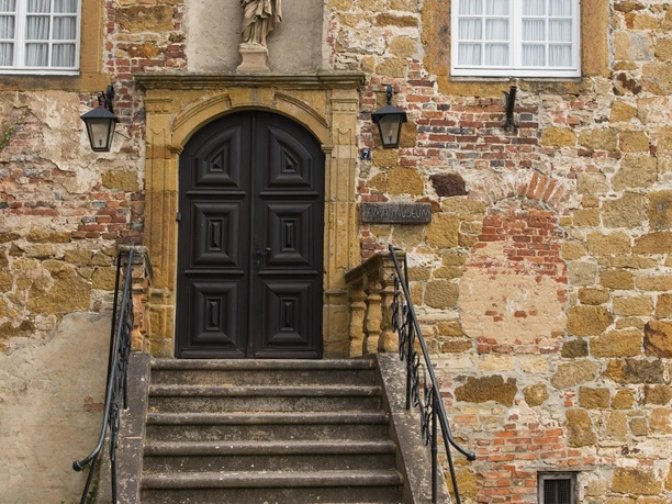 Historisches Gebäude aus Bruchstein mit Statue über schwarzem Türbogen, flankiert von zwei Laternen.