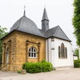 Die Loerdemann'sche Kapelle, ein Kleinod mitten in Herzebrock Kleine historische Kapelle mit schiefergedecktem Dach, flankiert von Bäumen und einem klaren Himmel.