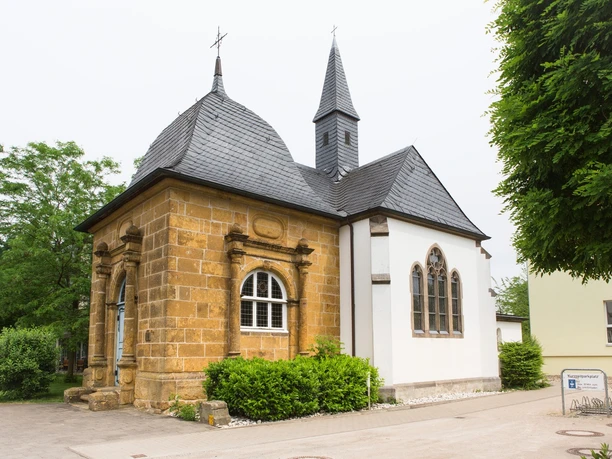 Die Loerdemann'sche Kapelle, ein Kleinod mitten in Herzebrock Kleine historische Kapelle mit schiefergedecktem Dach, flankiert von Bäumen und einem klaren Himmel.