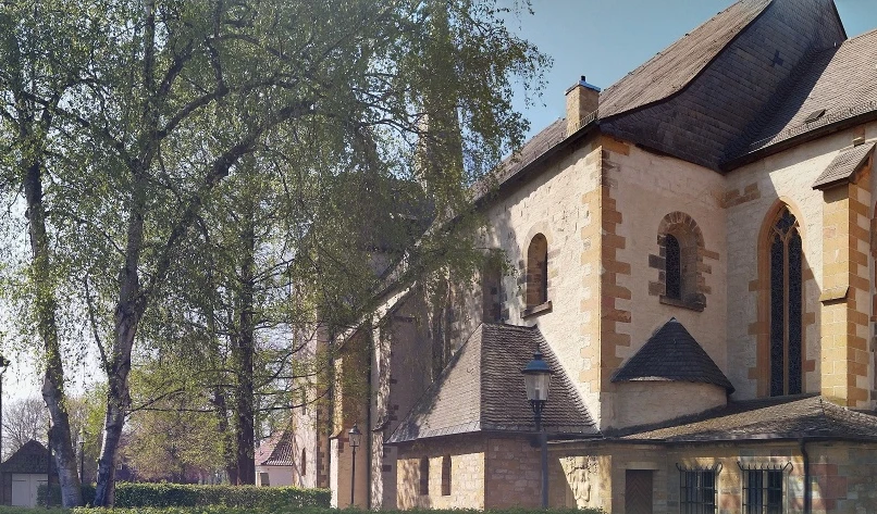 Historische Kirche mit romanischen Elementen neben belaubten Bäumen und angrenzenden Gebäuden.