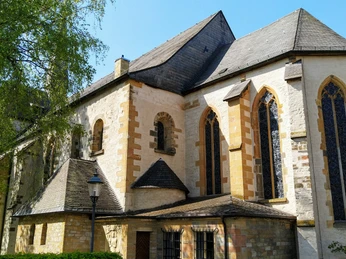 Historische Kirche mit gotischen Fenstern und schiefergedecktem Dach, umgeben von Bäumen und blauer Himmel.