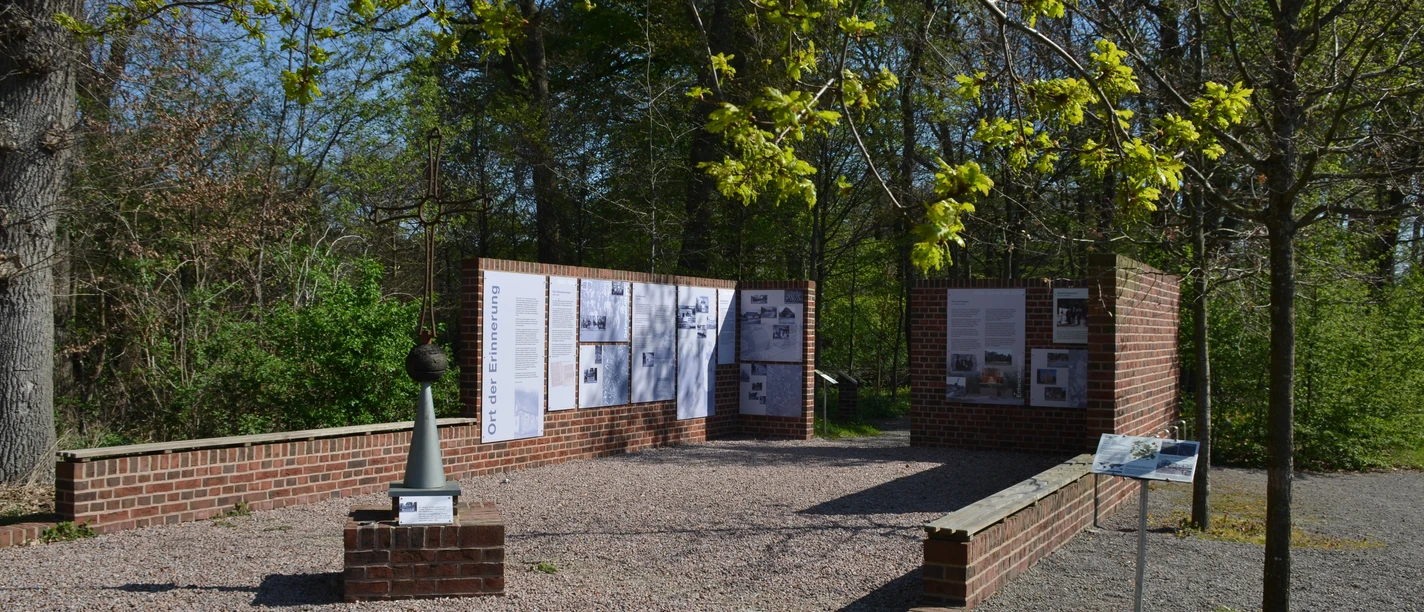 Notkapelle Wieste Gedenkstätte mit roten Backsteinwänden, Informationstafeln und Kreuz in grüner Parkumgebung.