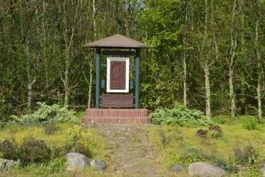 Hubertus-Denkmal Hubertus-Denkmal mit Holzrelief und Schrifttafel auf gemauertem Sockel vor grünem Waldrand.