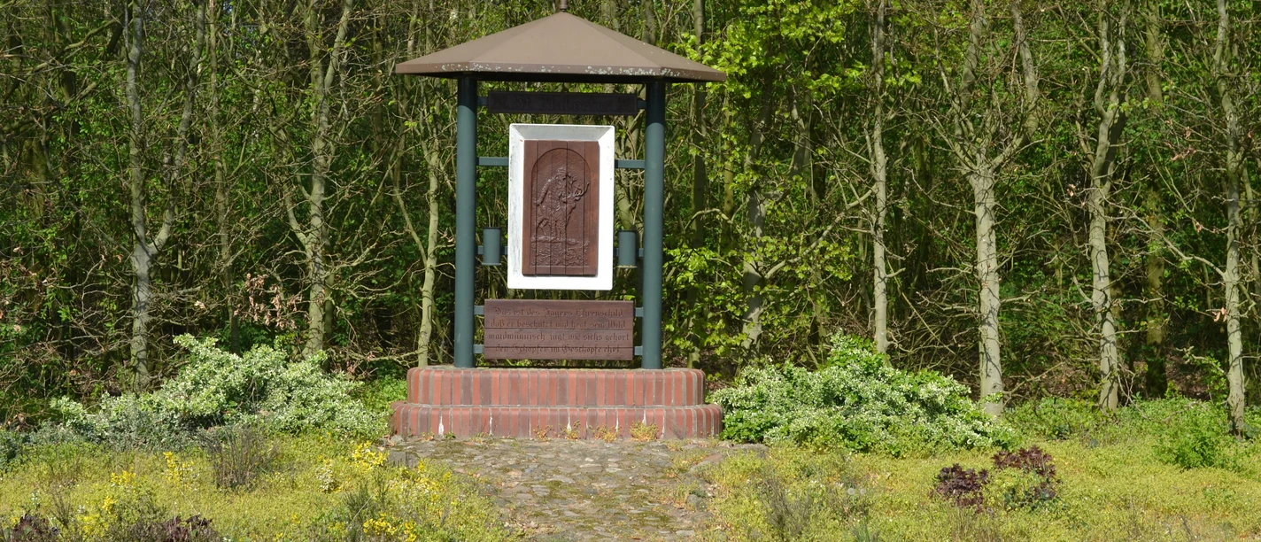 Hubertus-Denkmal Hubertus-Denkmal mit Holzrelief und Schrifttafel auf gemauertem Sockel vor grünem Waldrand.