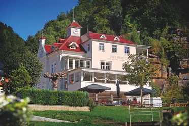 Villa Thusnelda met Café Bistro Daheim Een wit gebouw met een rood dak en een klokkentoren, omringd door bomen, huisvest Café Richter; er staan tafels met parasols voor.