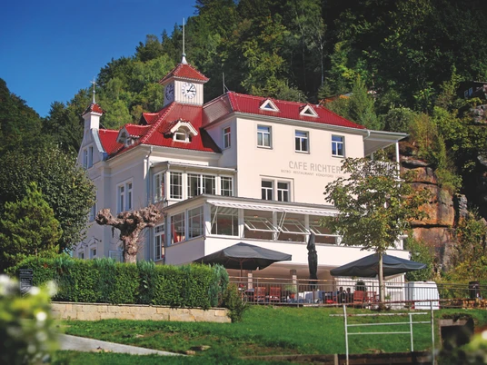 Villa Thusnelda s kavárnou Bistro Daheim Ein weißes Gebäude mit rotem Dach und Uhrturm, umgeben von Bäumen, beherbergt das Café Richter; davor stehen Tische mit Sonnenschirmen.A white building with a red roof and clock tower, surrounded by trees, houses Café Richter; tables with parasols stand in front of it.V bílé budově s červenou střechou a věží s hodinami, obklopené stromy, sídlí kavárna Café Richter; před ní stojí stolky se slunečníky.Biały budynek z czerwonym dachem i wieżą zegarową, otoczony drzewami, mieści kawiarnię Richter, przed którą stoją stoliki z parasolami.Een wit gebouw met een rood dak en een klokkentoren, omringd door bomen, huisvest Café Richter; er staan tafels met parasols voor.Un edificio bianco con tetto rosso e torre dell'orologio, circondato da alberi, ospita il Café Richter; davanti ad esso si trovano tavoli con ombrelloni.