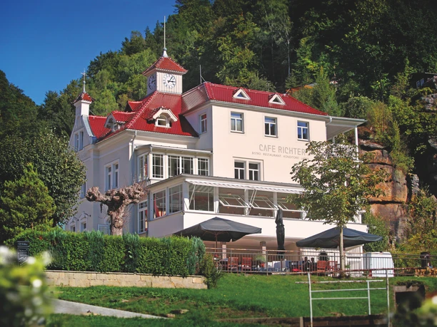 Villa Thusnelda with the Café Bistro Daheim A white building with a red roof and clock tower, surrounded by trees, houses Café Richter; tables with parasols stand in front of it.