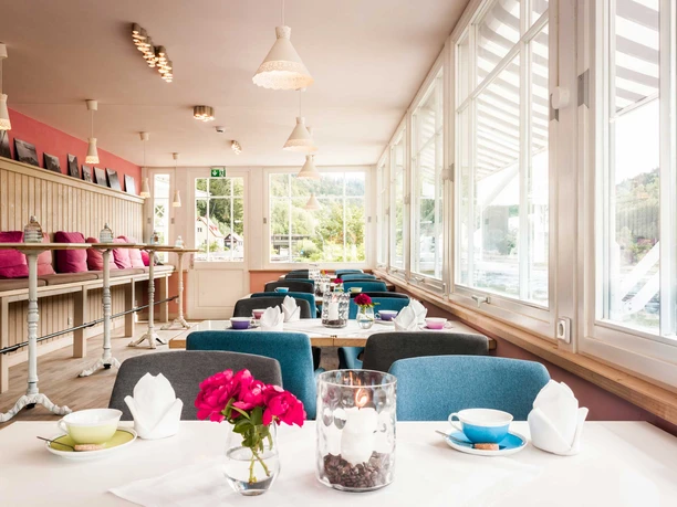 Café Bistro Daheim Interior Bright, cozy café room with large windows, blue chairs, pink cushions and decorative flowers on the tables.