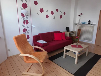 Wohnzi, FeWo Krauß Ein Wohnzimmer mit rotem Sofa, orangenem Sessel, Holztisch, Blumenmuster an der Wand und Holzboden.