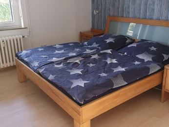 1. Schlazi, FeWo Krauß Schlafzimmer mit Doppelbett, blauer Bettwäsche mit Sternenmuster, hellem Holzboden und Fenster.